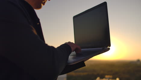 A-programmer-with-a-laptop-sits-on-the-roof-and-writes-code-at-sunset.-Remote-work-freelancer.-Freedom-to-work.-Typing-on-a-keyboard-at-sunset-with-a-view-of-the-city.